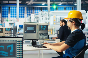 Factory worker wearing a yellow hard hat sits at a computer station, monitoring production dashboards on multiple monitors in a factory.